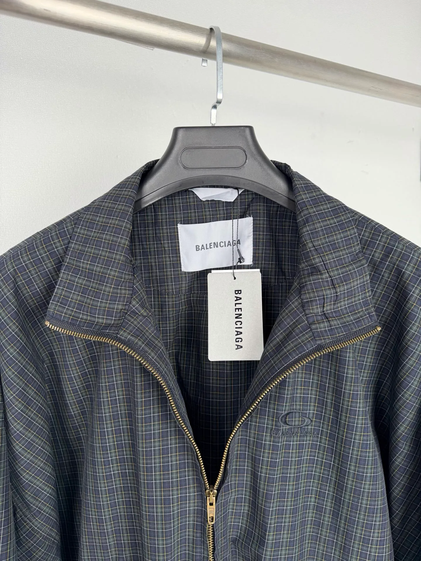 BALENCIAGA/balenciaga SS 26 two-way zipper opening and closing embroidered plaid jacket is based on the full-area no-dead-corner grid technology as the technical core, superimposed on multiple difficult processes such as two-way zipper composite structure, positioning heavy embroidery, and full-cut symmetrical sewing. The complexity of the process and the scrap rate are far higher than that of conventional garments. It is a ceiling-level presentation of street luxury craftsmanship. This product is made of custom-made yarn-dyed plaid original fabric. It needs to complete four pre-processing processes of ultra-dense pre-shrinking, weft density correction, tension leveling and flat-width shaping before being put into production. The shrinkage error of the fabric's longitude and latitude is strictly controlled within 0.2%, which completely eliminates the hidden dangers of plaid deformation and alignment deviation after the garment is worn, laying an irreplaceable foundation for the whole piece to align. The pre-processing time of a single fabric is three times that of an ordinary jacket. The core process difficulty is the full-area through-alignment technology, which requires all exposed pieces such as the front and rear panels of the body, large and small sleeve panels, lapel collars, pocket flap welts, placket stops, and side seam panels to achieve horizontal penetration of horizontal grids, vertical alignment of vertical grids, and complete mirror symmetry of left and right panels. The alignment tolerance is strictly controlled within 0.1mm, and the industry standard is only 0.5mm. Cutting pieces are arranged using a zero-loss precise positioning method. The loss rate of a single fabric is increased by 20%. Any deviation in the pattern of the cutting pieces is judged as a defective product. The experience and precision requirements of the cutting masters have reached a strict level. At the structural level, it is equipped with a two-way double-head customized zipper opening and closing system. The zipper tooth chain and placket pattern must be 100% aligned in the center seam. When the zipper is installed, the plaid alignment and stop smoothness are simultaneously completed to prevent the plaid from being pulled or skewed due to zipper installation. The front loop sportsicon heavy embroidery adopts positioning embedded technology. The embroidery stitches need to accurately follow the warp and weft direction of the check pattern. The positioning error of the embroidery frame does not exceed 0.05mm. The floating embroidery process avoids the stitch tension from pulling the fabric texture and achieves the perfect symbiosis between the embroidery pattern and the check texture. The entire garment adopts segmented alignment sewing and full-process quality inspection and control. From cut pieces to finished products, it needs to undergo 7 alignment checks. Every seam, structural line, and decorative piece serves the integrity of the pattern. The complexity of the process, labor cost, and quality control standards are all benchmarked against the high-end garment level. It has both pioneering design and unrepeatable ingenious workmanship. Size: 46 48 50 52